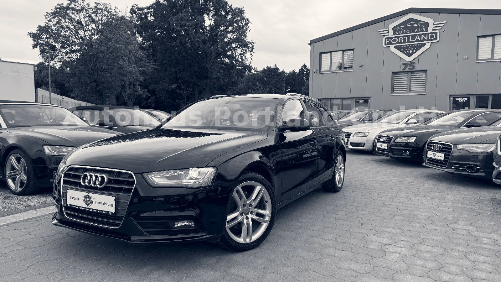 Schwarzer Audi A4 Avant auf dem Hof von Autohaus Portland in Hannover-Misburg, im Hintergrund das Firmengebäude mit Logo